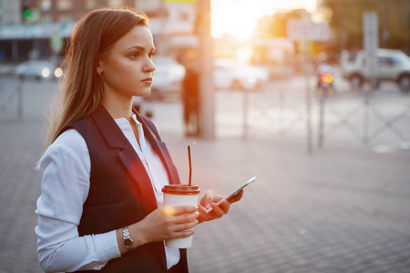 年轻漂亮的女人，带着智能手机和纸咖啡杯，在夕阳的背景下，在城市街道上。复古色调过滤效果颜色风格。