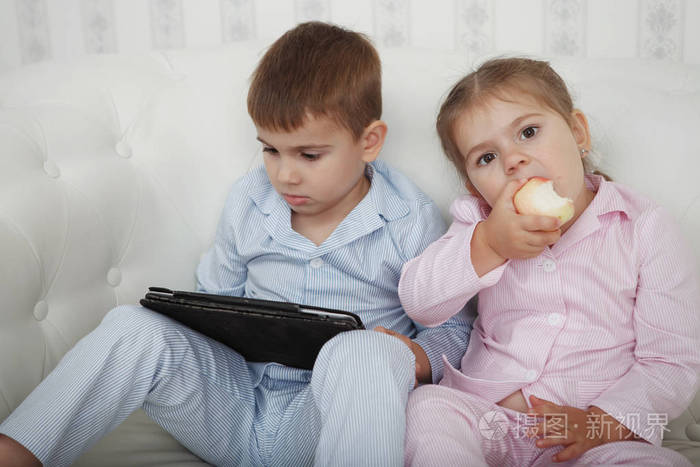 兄妹俩穿着粉色和蓝色睡衣坐在白色沙发上，看着平板电脑。