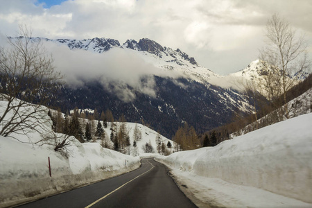 空的扭曲的沥青道路延伸到深雪，走向美丽的明亮的白色阿尔卑斯山，陡峭的岩石木质雪山峰下多云的冬季天空。 寒冷晴天的冬季景观。