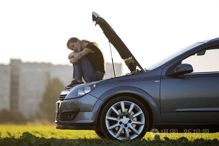 年轻迷人的悲伤女人坐在车上绿色的草地上模糊的公寓楼和晴朗的天空