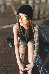 与纹身和长发绺在蓝色帽子的年轻女孩坐在长板的背景下混凝土结构图片