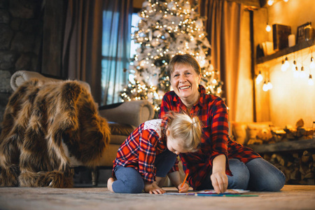 祖母和孙女坐在树前的地毯上画画。 家庭圣诞概念。