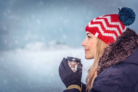 一位美丽的女士戴着时尚温暖的帽子，手里拿着一杯热咖啡，享受着下雪的日子，在寒冷的冬天在户外玩得很开心。