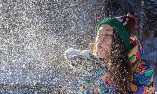 美丽的冬天女孩吹雪在寒冷的冬季公园。户外。飞行雪花。晴天。背光。快乐的美女年轻女子在冬季公园玩耍