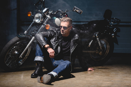 戴墨镜的帅哥骑车坐在车库的摩托车旁边的地板上