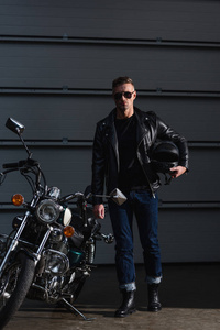 穿着黑色太阳镜和皮夹克的时髦自行车手，站在摩托车旁边，戴着头盔