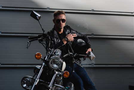 穿着黑色太阳镜和皮夹克的帅哥坐在车库里的摩托车上