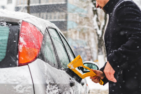 在下雪时，穿着黑色外套的年轻人用黄色刷子清洁他的车。 冬天恶劣的天气。