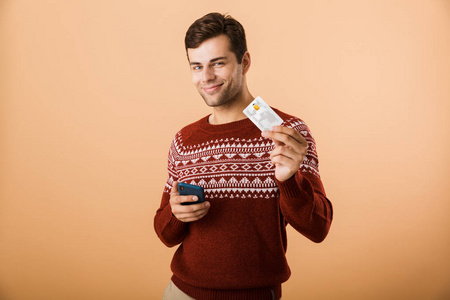 一位穿着毛衣的年轻人，站在米色背景下，手里拿着显示信用卡的手机