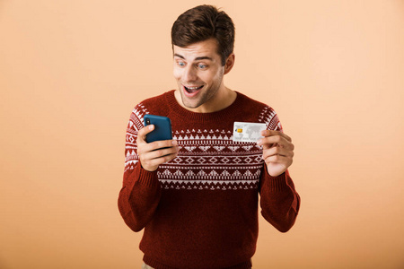 一位穿着毛衣的开朗的年轻人站在米色背景下，手持显示信用卡的手机
