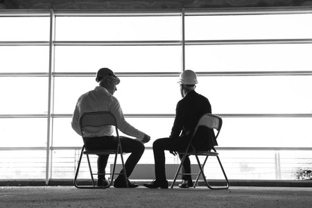 男子工程师建筑商在桌旁工作, 在建筑工地上工作, 进行商务谈判。在窗口的背景下的剪影