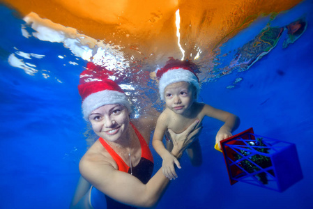 微笑的母亲和一个婴儿在游泳池里游泳，戴着蓝色背景的圣诞老人的红色帽子。 孩子手里拿着一个圣诞玩具。 肖像。 图像的水平方向。