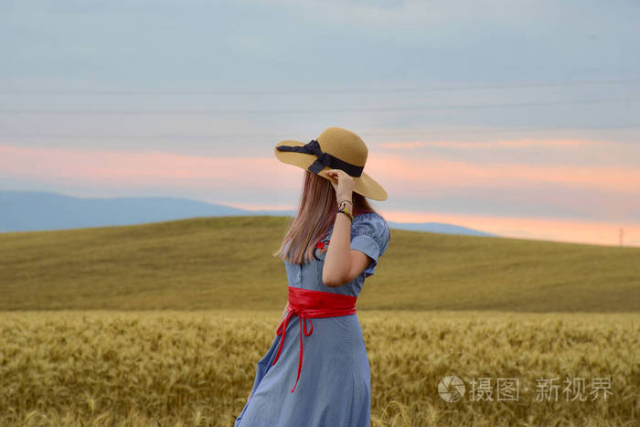一个穿着蓝色连衣裙和一顶浅色帽子的女孩站在麦田的中间,黄色的