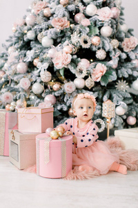 可爱的小女孩穿着bklom连衣裙坐在面粉里，打开一个盒子，里面有一个背景圣诞树的礼物。 圣诞节假期概念