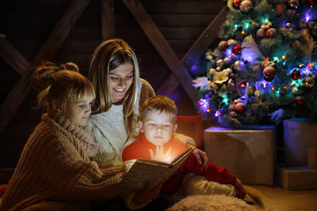 圣诞快乐, 新年快乐。美丽的家庭在圣诞节的内部。漂亮的年轻母亲在圣诞树附近给女儿和儿子读书