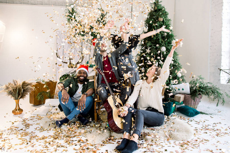 圣诞节假期和新年聚会的概念。 四个多种族的朋友庆祝圣诞节在舒适装饰的客厅圣诞树。 彩纸彩片