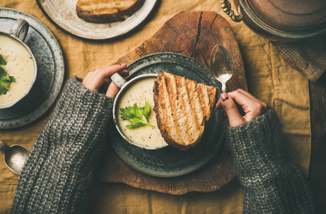秋季或冬季家庭晚餐。 平躺秋暖芹菜奶油汤和女性手与勺子在黄色亚麻桌布上的顶部视图。 舒适食品，素食，健康和缓慢的食物概念