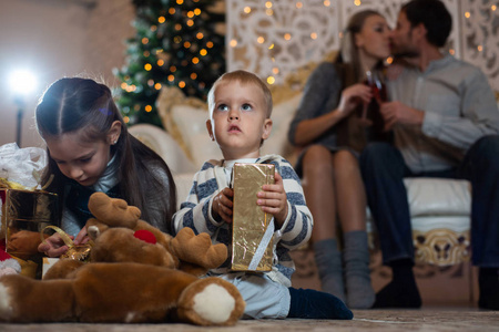 小哥哥小姐姐拿着礼物坐在地上，背着父母和圣诞树