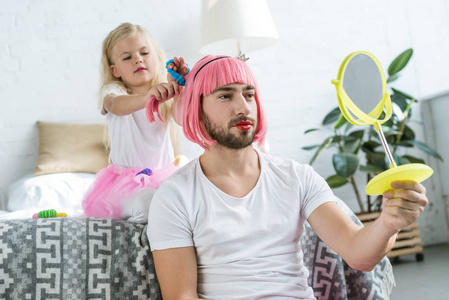 可爱的小女儿，穿着图坦裙子，和戴着粉红色假发的父亲玩，看着镜子