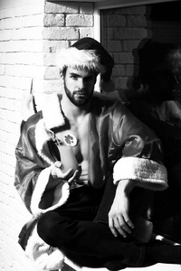 年轻英俊具髯毛具肌肉发达的上身，坐在窗台上和含酒精的鸡尾酒玻璃坚持黑和砖的背景墙上红色的圣诞老人帽子和外套圣诞男
