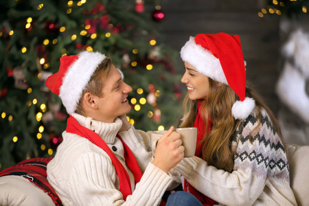 可爱的年轻夫妇戴着圣诞帽，带着热巧克力在家庆祝圣诞节