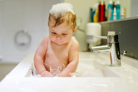 可爱可爱的宝宝，考虑在洗涤池浴和抢水龙头。蓝色的大眼睛，开心和玩肥皂泡沫的小健康女孩