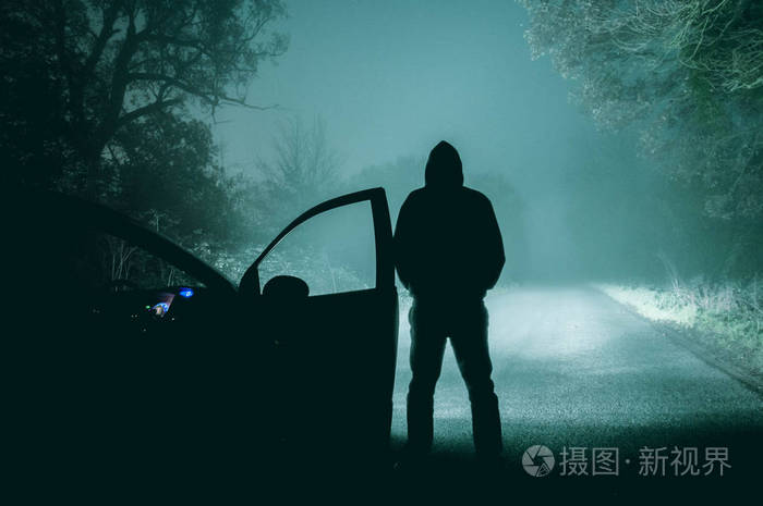一个孤独的,头戴头套的身影站在一辆汽车旁边, 看着一辆空着的人