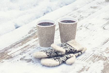 在冬天，两个纸工艺品玻璃杯和一杯热饮料咖啡可可覆盖葡萄酒和白色和黑色手套在户外的桌子上。 冬季时间概念。 沐浴在寒冷中。