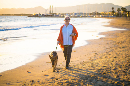 美丽的退休老年妇女和宠物德国舍帕德狗走在海岸海洋海滩上的友谊福利动物保持积极的退休生活方式和狗友好的旅游。