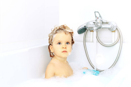 可爱的可爱宝宝在水龙头背景下的洗手池洗澡。 健康的小女孩，有着蓝色的大眼睛，玩着肥皂泡沫。 认真地抬起头来。 手中的玩具
