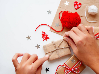 女人正在用手工纸包装圣诞节和新年DIY礼物。 用白色和红色线捆绑的礼物。 白色背景上的盒子和星星纸屑。