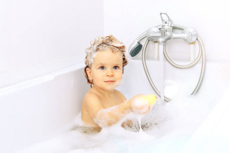 可爱的微笑宝宝在浴室里用白色背景的黄色橡胶鸭洗澡。泡沫和气泡环绕..湿肥皂毛。蓝色的大眼睛
