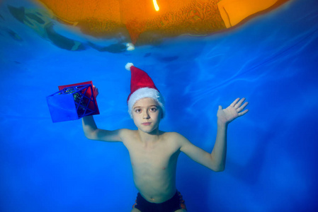 一个小男孩穿着红色的圣诞帽在游泳池一顶红色的圣诞帽，手里拿着一件蓝色背景的礼物，看着相机。 肖像。 概念。 景观定位的形象。