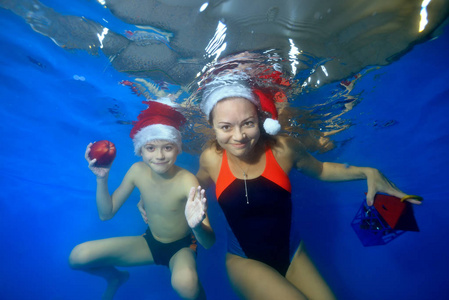 幸福的家庭母亲和孩子在游泳池里的蓝色背景上，穿着红色的圣诞帽，在水下为相机做运动和姿势。 肖像。 概念。