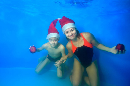 一个带着微笑母亲的小男孩游泳，在池底的摄像机前摆姿势，戴着蓝色背景的红色圣诞帽，手里拿着红色的苹果。