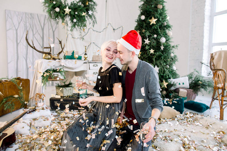 年轻时尚的情侣们坐在圣诞树附近的地板上, 扔着金色的五彩纸屑, 庆祝新年, 快乐的聚会, 开心, 面带微笑