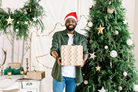 非洲裔美国人穿着时尚的绿色衬衫, 在圣诞树的背景下, 带着礼品盒, 戴着圣诞老人的帽子, 露出了灿烂的笑容