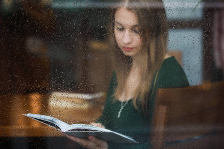 妇女在咖啡馆看书, 在雨天透过潮湿的窗户看风景