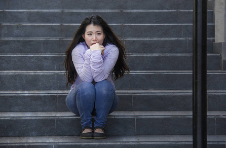 年轻的悲伤和沮丧的亚洲学生妇女或被欺负的青少年坐在户外的街道楼梯上，不知所措，焦虑地感到绝望，患有抑郁症