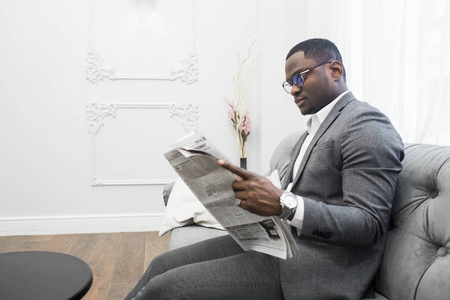 年轻的非洲裔美国商人穿着灰色西装坐在沙发上看报纸