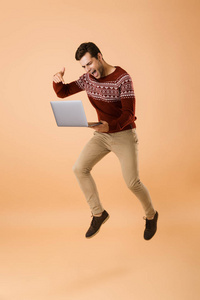 一位穿着毛衣的年轻人用手提电脑在米色背景下跳起来，心情激动