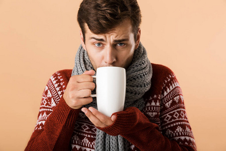 一个穿着毛衣和围巾的迷人的年轻人从米色背景的杯子里喝茶