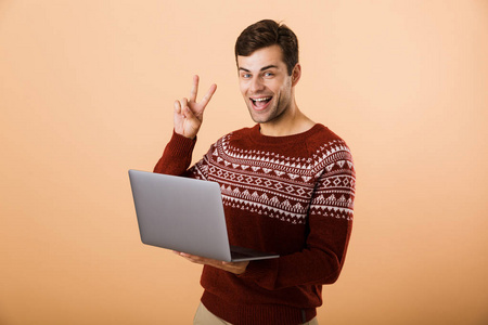 描绘了一个穿着毛衣的年轻人，用笔记本电脑在米色背景下孤立地站着，表现出和平的姿态