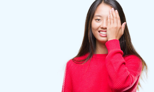 年轻的亚洲女人穿着冬天的毛衣，在孤立的背景上覆盖一只眼睛，脸上带着自信的微笑和惊讶的情绪。