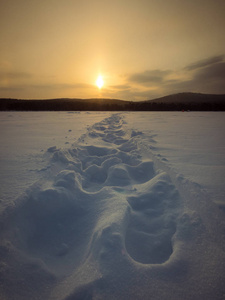 新靴子脚印在雪上的湖面上，冰在夕阳的灯光下，在冬天的晚上。 背景。