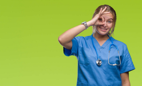 年轻的高加索医生女人穿着外科医生制服，在孤立的背景上做着OK的手势，用手微笑的眼睛透过手指看着幸福的脸。