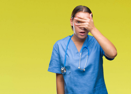 年轻的白种人医生女人穿着医用制服，在孤立的背景上偷看，震惊地遮住脸和眼睛，用手透过手指看着尴尬的表情。