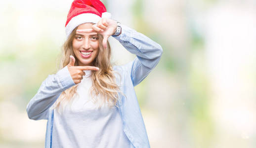 美丽的年轻金发女人戴着圣诞帽，在孤立的背景上微笑，用手和手指制作框架，用快乐的脸。 创造力和摄影理念。