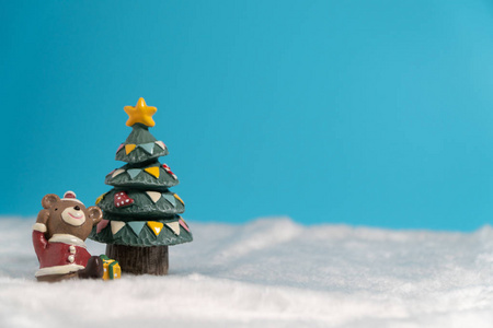 快乐的棕熊穿着圣诞老人礼服，带着礼品盒坐在圣诞树附近的雪地上。