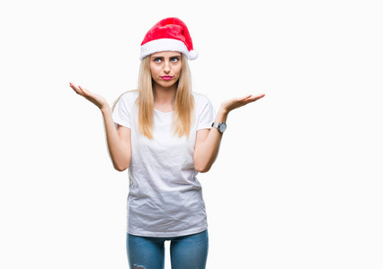 年轻漂亮的金发女人圣诞帽在孤立的背景上毫无头绪，表情混乱，举起手臂和手。 怀疑概念。